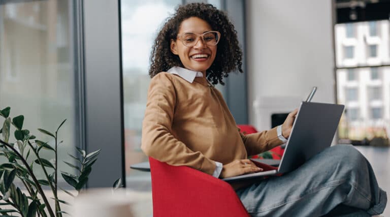 a women sitting happily in her red chair with her phone in her one hand and her laptop on her lap