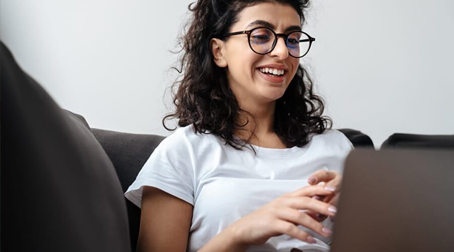 A dark haired curly women wearing her black glasses sitting in front of her laptop
